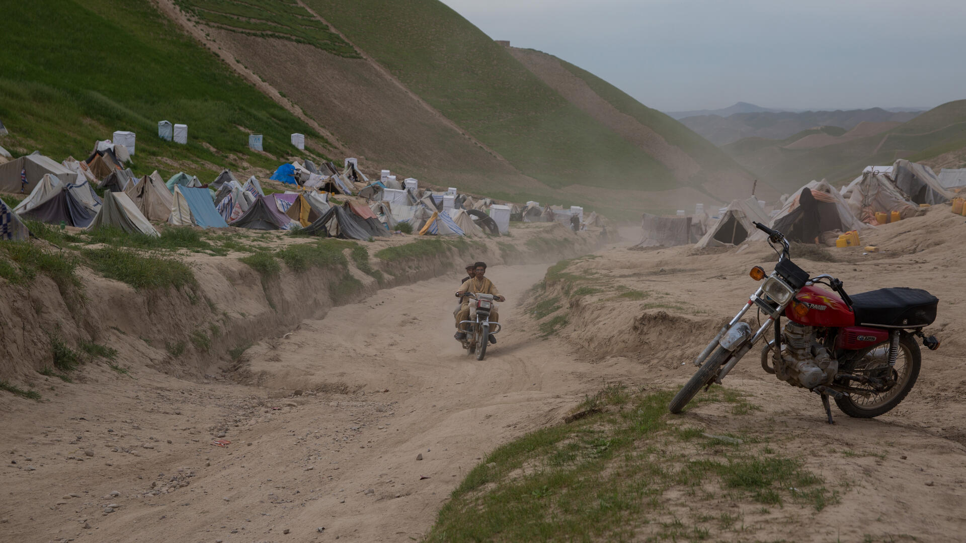 A camp for drought displaced people in Afghanistan. there is a motorcycle in the foreground as well as two men on a motorcycle on the road, and tents in the background on a mountainous landscape.
