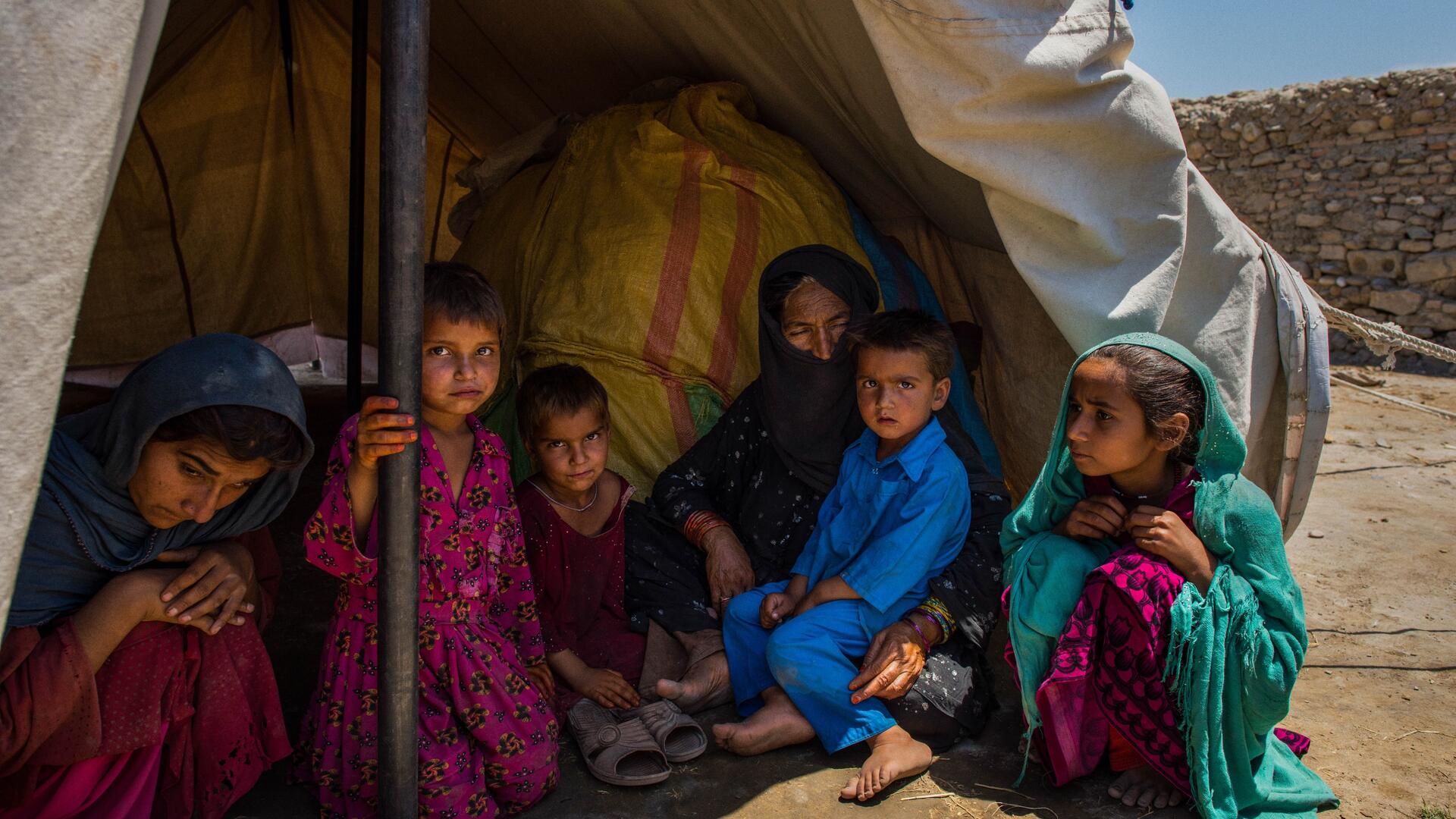 Women and children sit in the shade of one of their family's tents in a displacement camp in a desert landscape in Afghanistan..