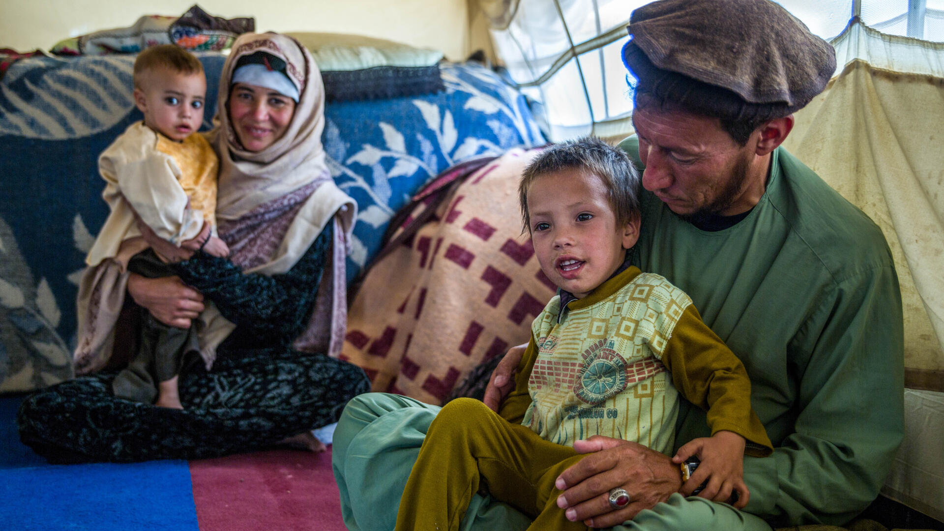 A farming family in Afghanistan who had to flee their home due to drought. Cash they received from the IRC allowed them to take their child to the hospital. A mother and father in Afghanistan sit on the ground among blankets and pillows, each with a child on their lap. The boy in the front is smiling and his father is looking at him.