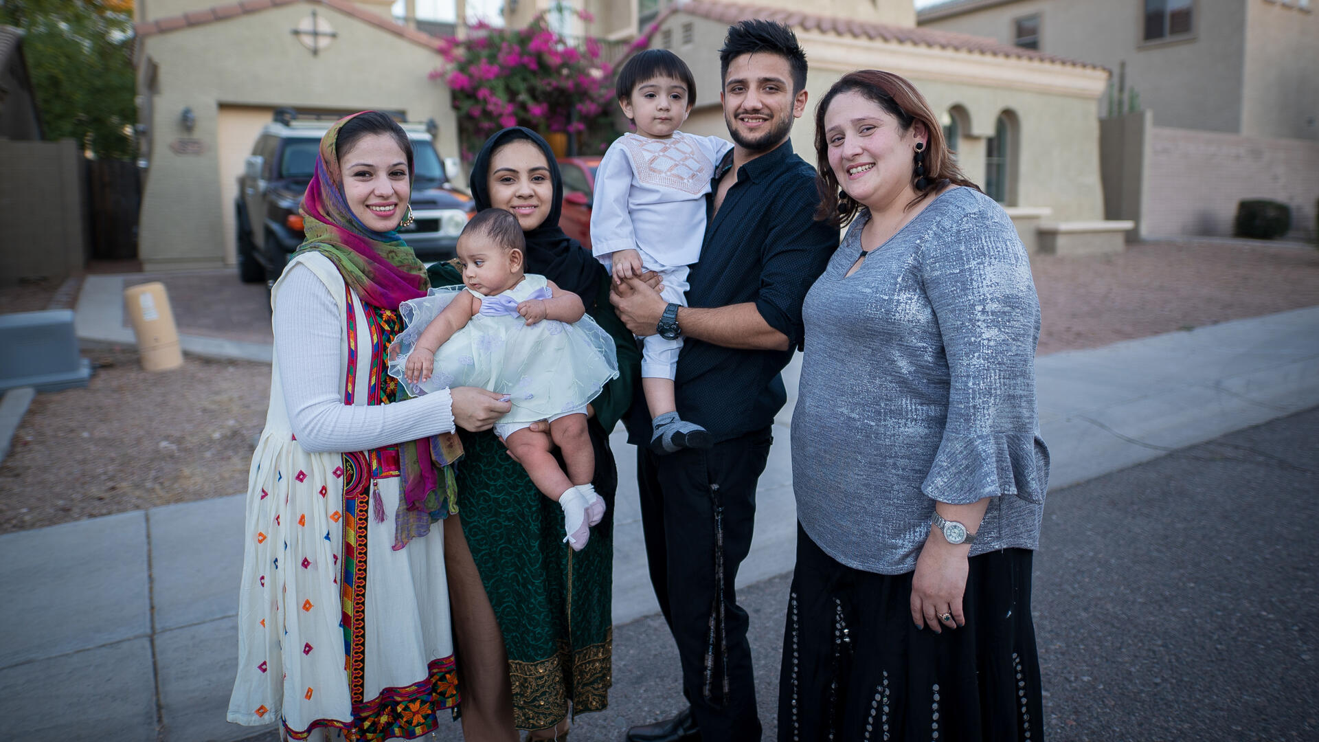 Muska, an Afghan refugee, with her family in Phoenix. With the help of the IRC, Muska started a business selling imported traditional Afghan and Pakistani clothes. She is also a pre-med student. Muska, a woman in her twenties, stands in front of her home with three adult family members, a toddler and a baby.