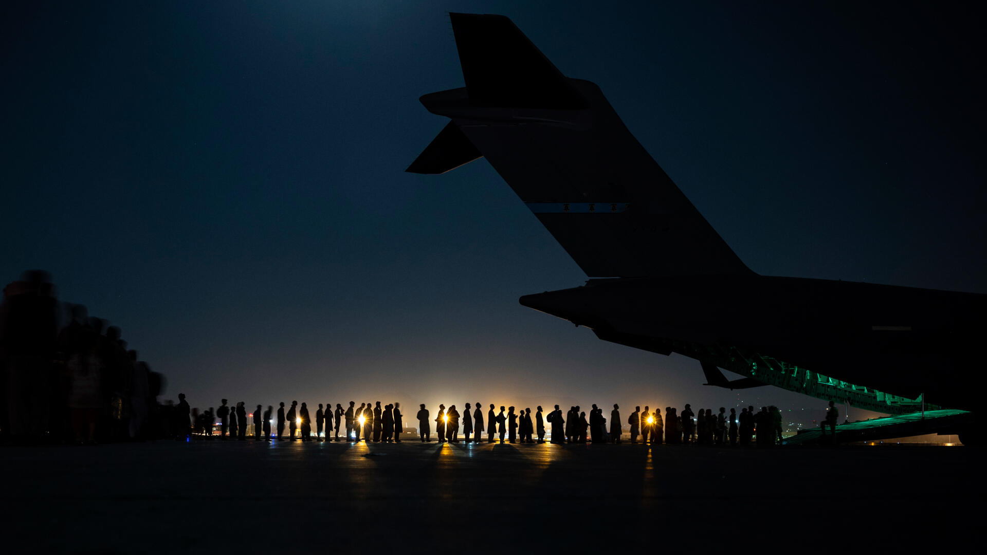 An airplane prepares to load evacuees at Hamid Karzai International Airport. A night shot, with the tail of an airplane in the foreground and a line of people in the background. We can see a light in the distance and just the silhouette of the plane and people.