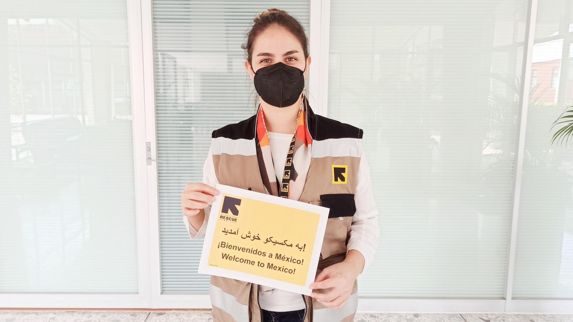Fatima, a home visiting officer for the IRC in Mexico, holds a sign that says "welcome to Mexico" in English, Spanish and Dari. She is wearing a mask and an IRC vest. 