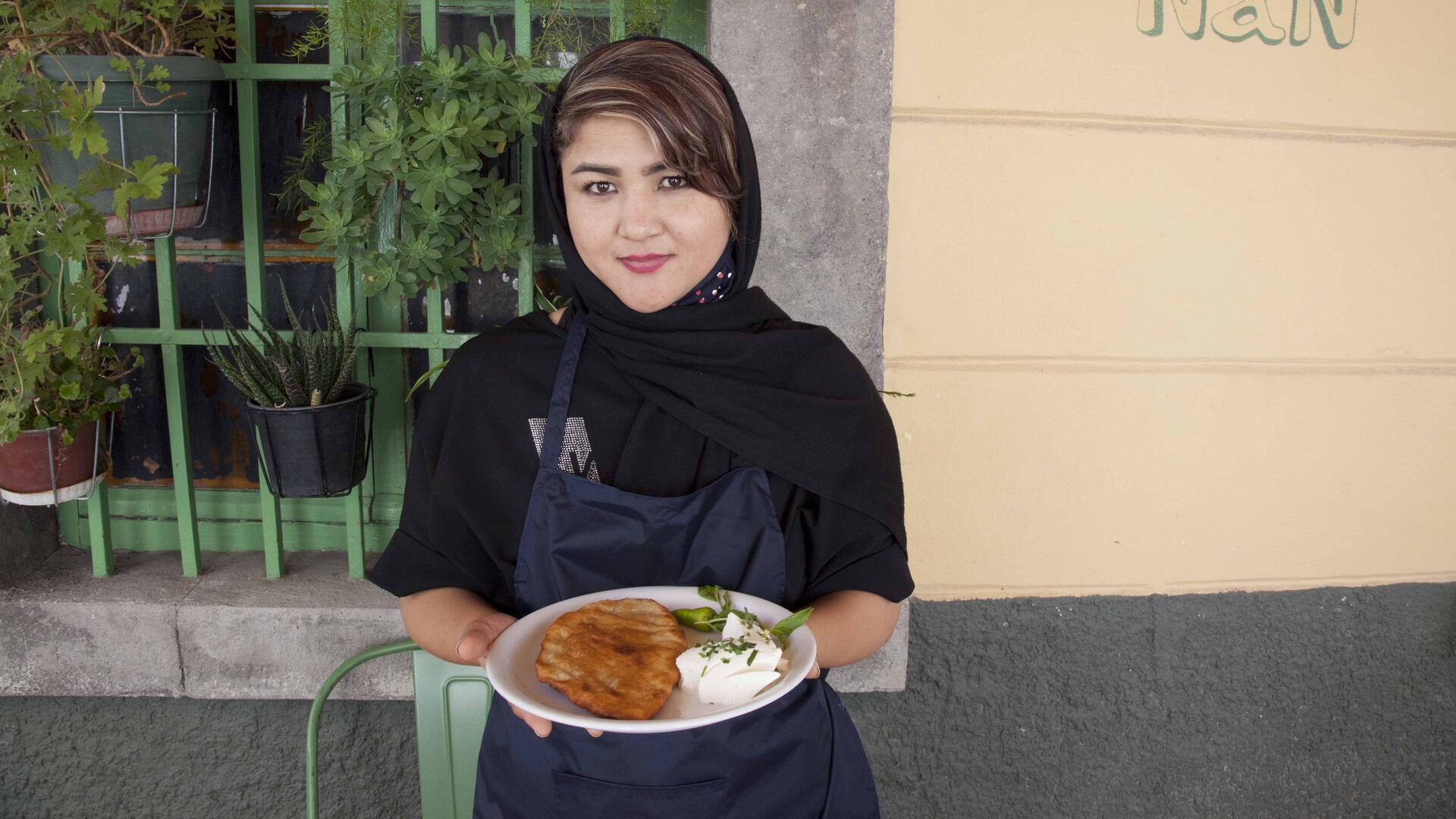 Hafeza, a refugee from Afghanistan living on the Greek island of Lesbos, holds her homemade Afghan Bolani Wearing an apron, Hafeza stands in front of a building holding a plate of Afghan Bolani.