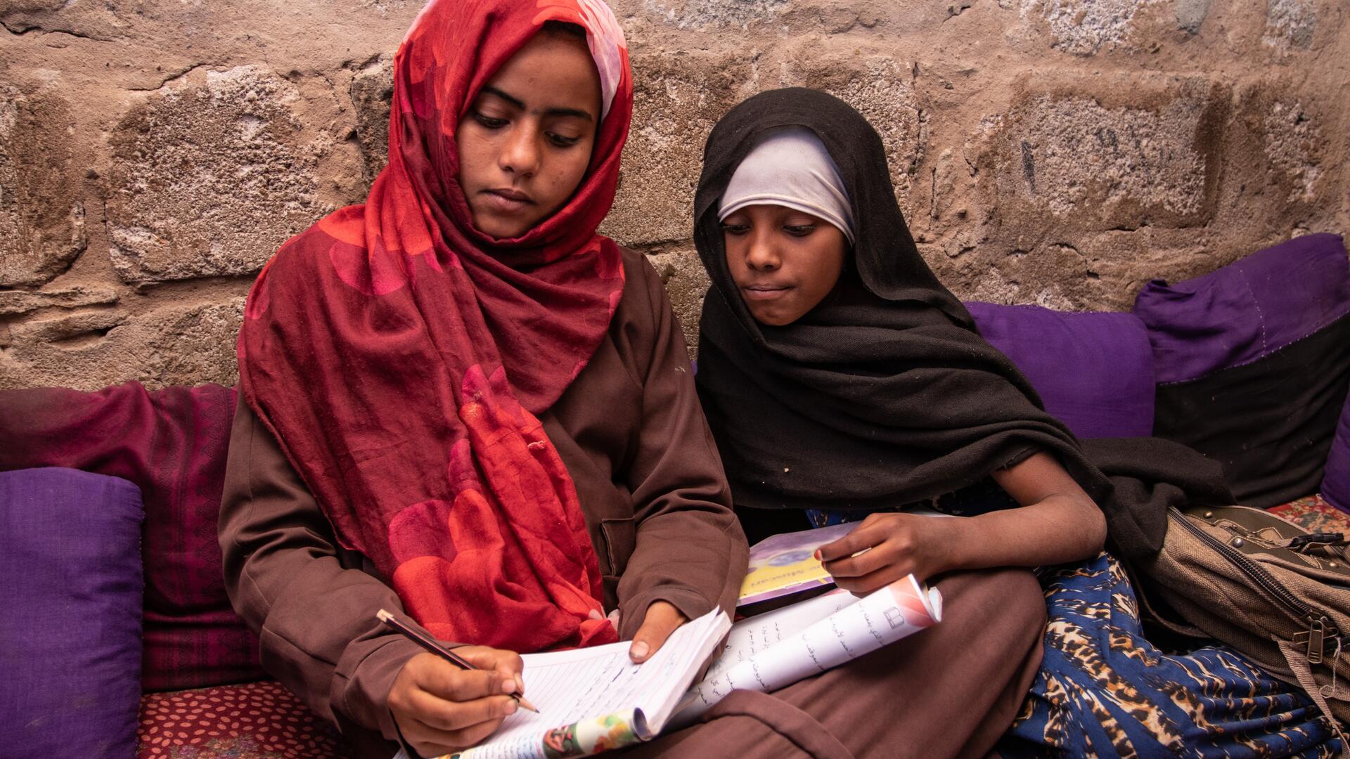 Na'aem, 11, and her best friend Aisha, 10, sit together on the ground studying a school book in a displacement camp in Yemen.. 