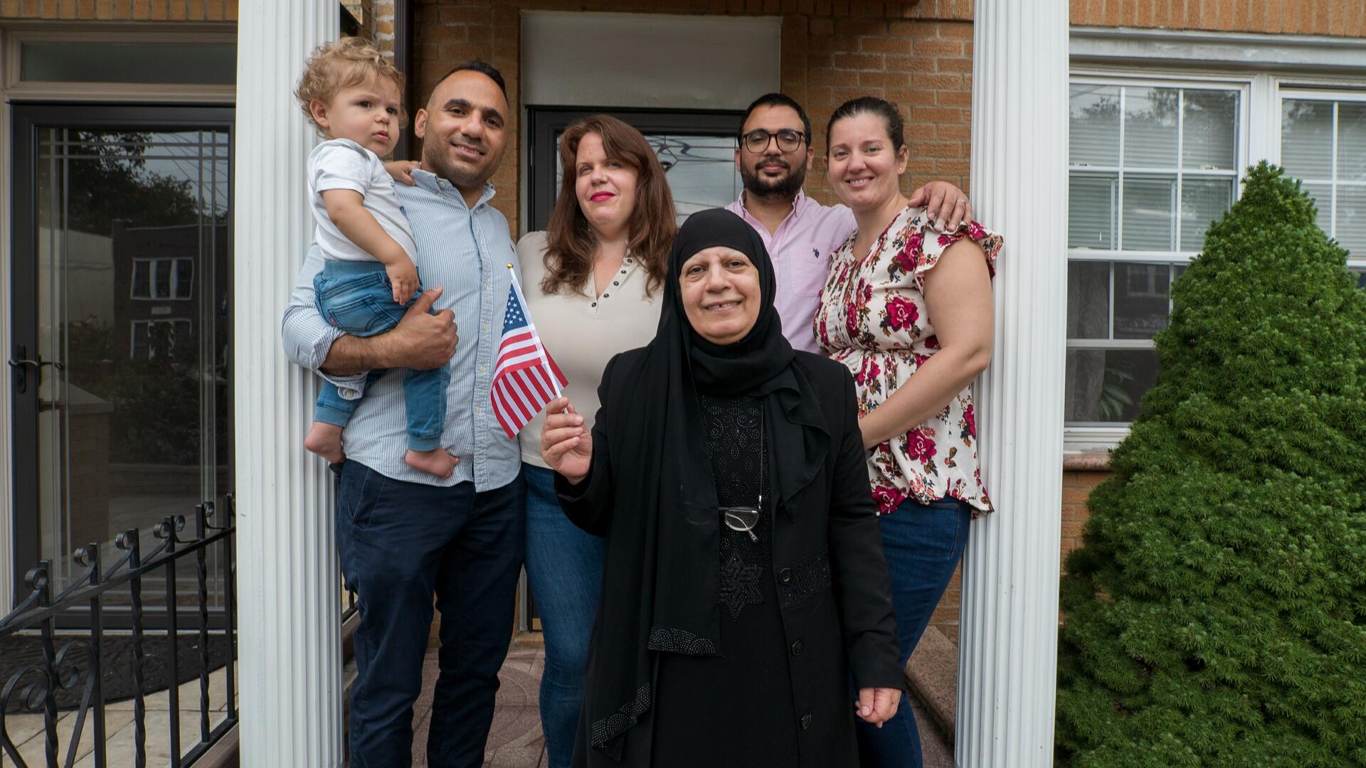 Maha al-Obaidi, a 66-year-old Iraqi woman, stands holding an American flag with her family on the porch of their home in New York City.