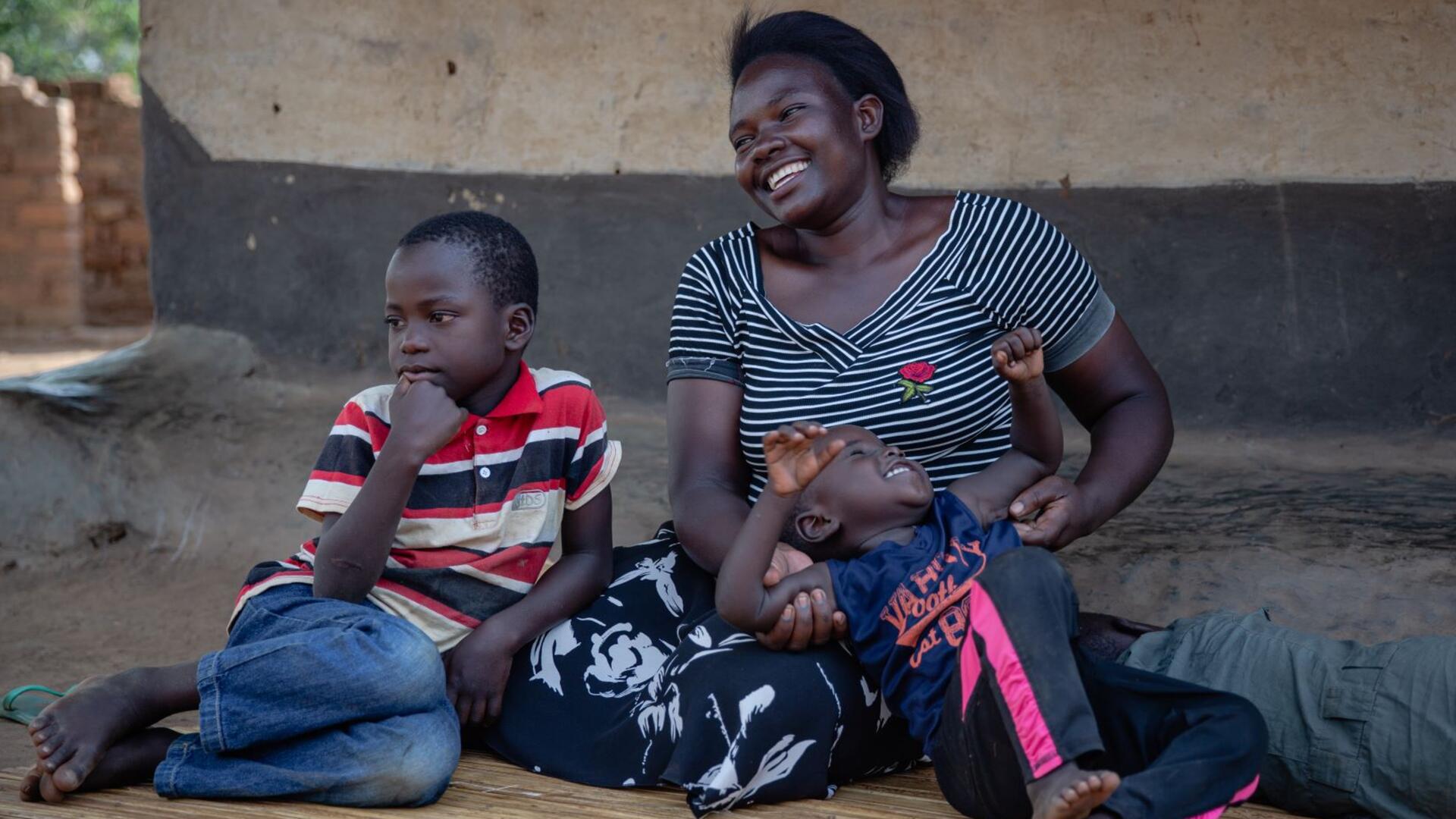 Jackie with her children Jackie sits on the ground smiling with her two sons. The older is leaning against her and looking into the distance and the younger is lying on his back on his mothers lap, laughing.