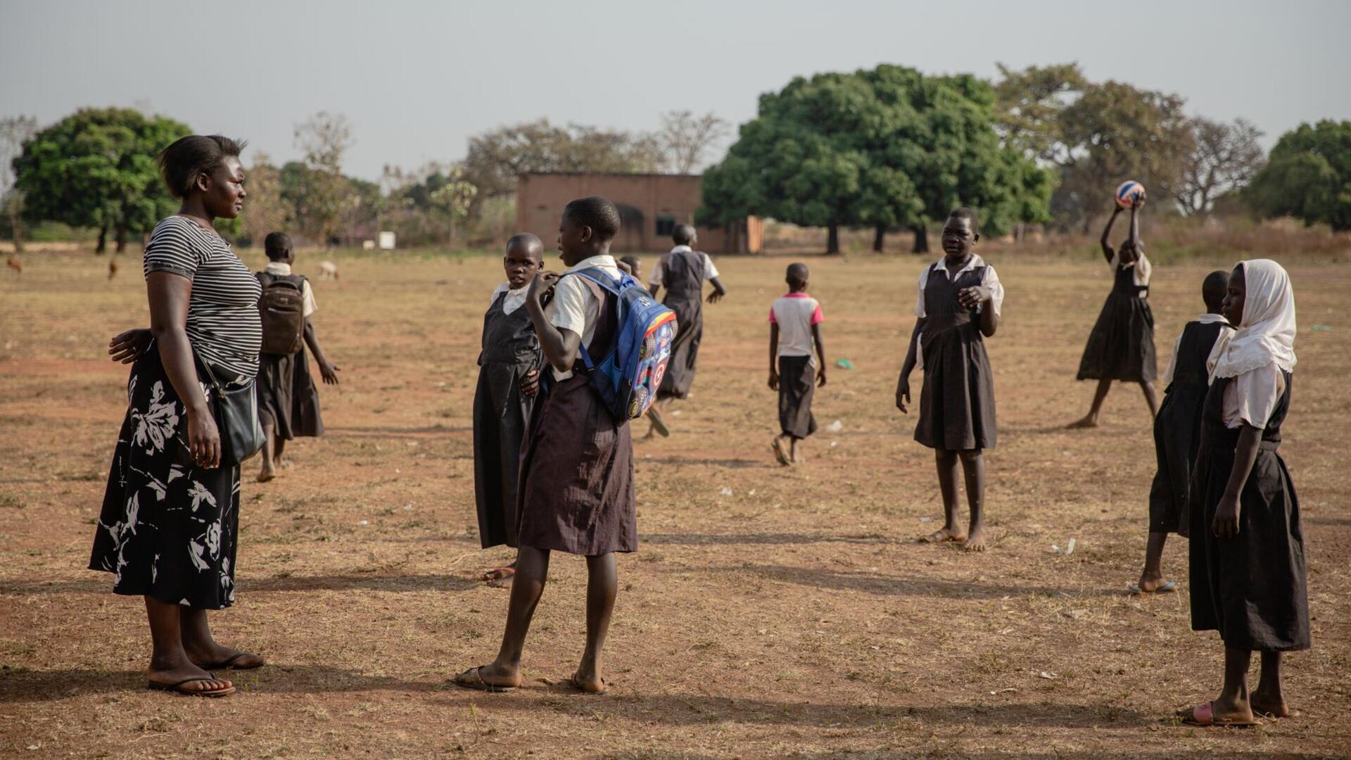 Jackie at work with Logoa Women Jackie stands in a field with young girls in school uniforms. One holds a ball as they prepare to play netball.