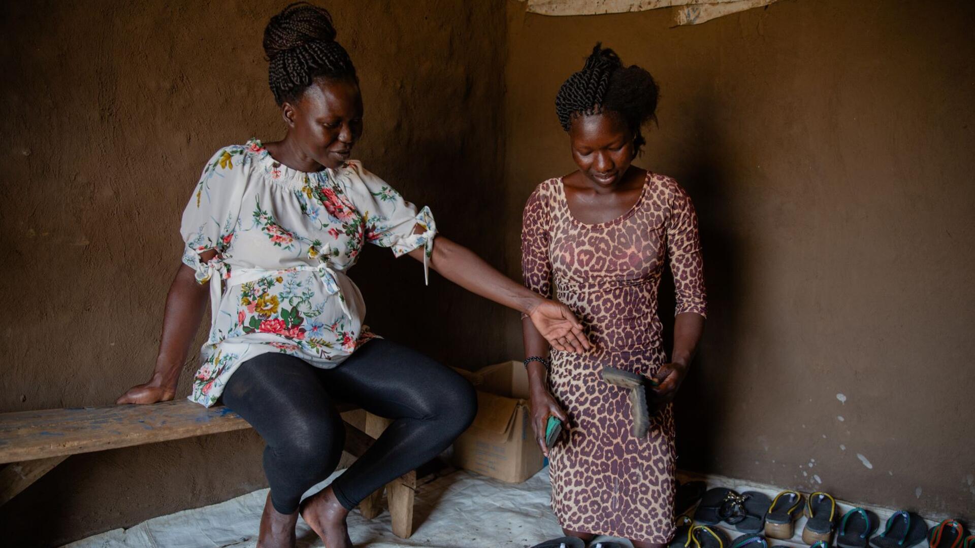 Grace works with refugee Vivian to help her start her own business Grace sits on a bench next to a refugee named Vivian, who is kneeling next to shoes arranged neatly on the ground. She is holding one of the shoes and looking down at them.