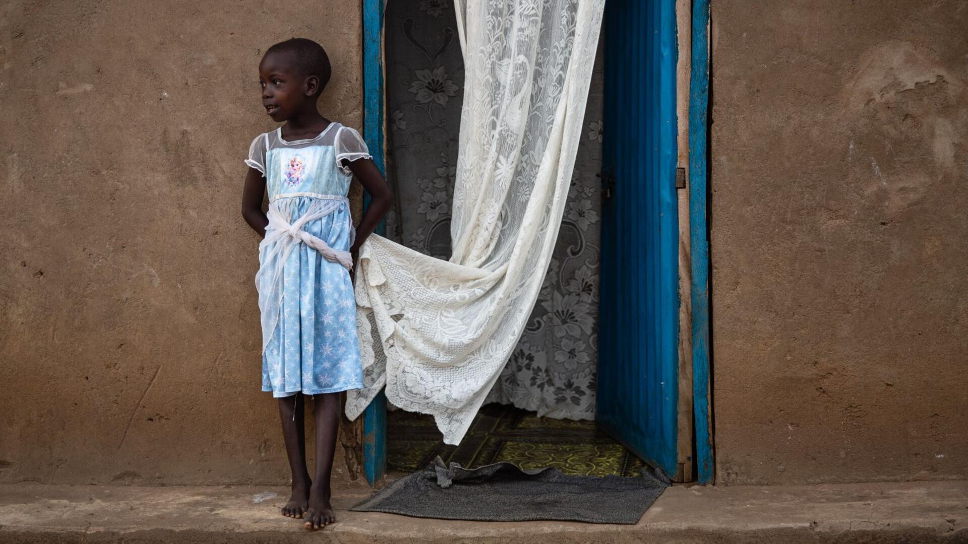 Foni Grace’s six-year-old daughter, Nancy, stands outside their home. She is looking to the side and holding the edge of a white curtain. 