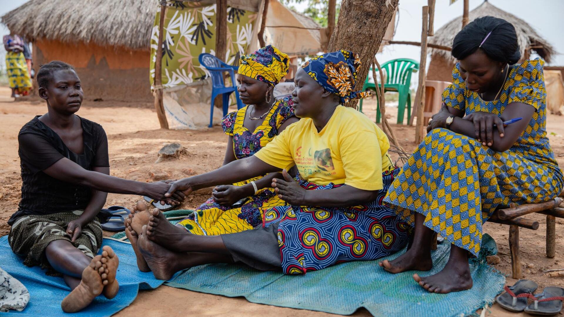 Jemimah and other Togoleta members counsel a woman named Cecilia. Jemimah is sitting on a blanket on the ground next to Cecilia, holding her hand. The other two women are sitting on chairs nearby. 