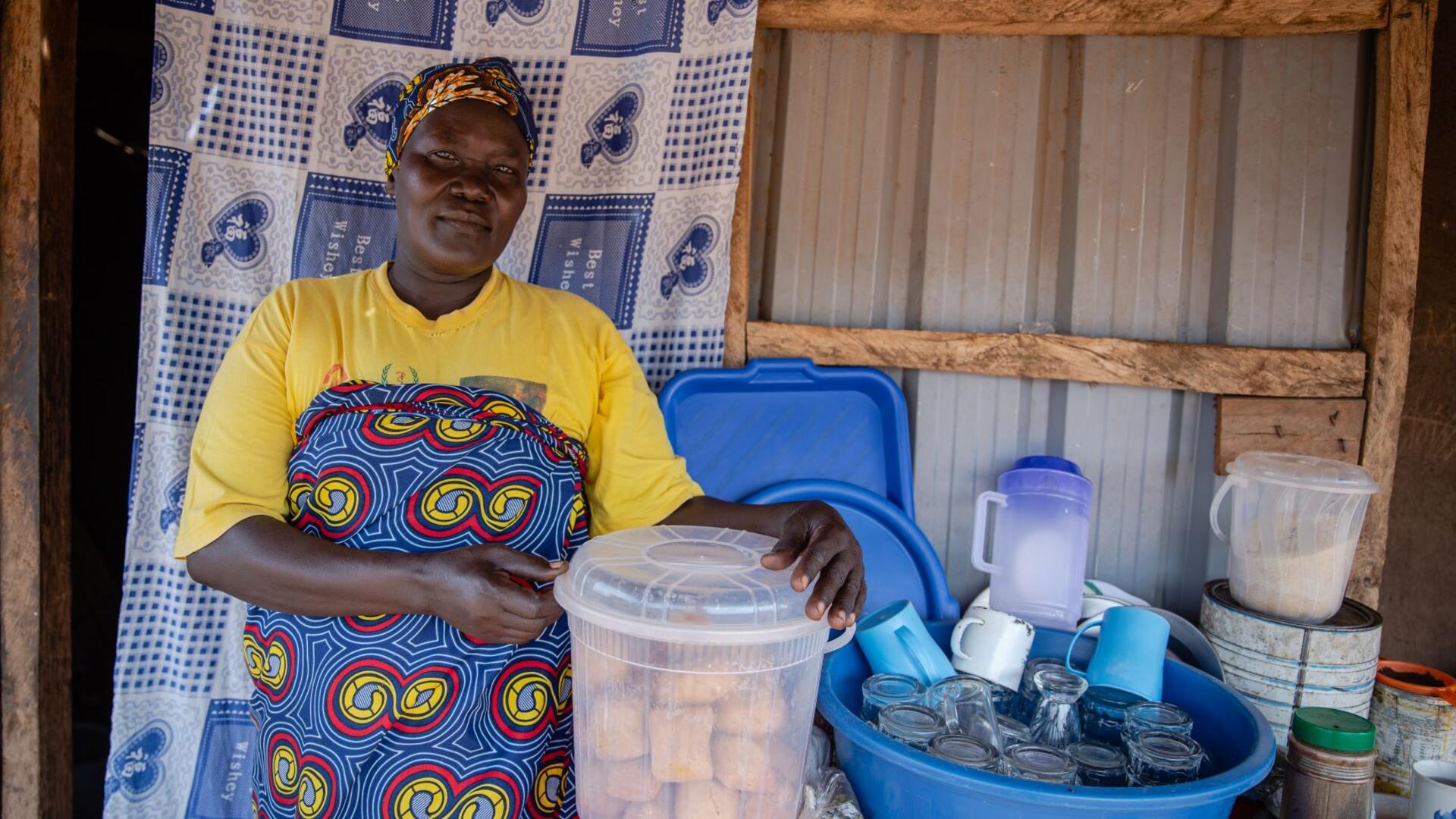 Jemimah poses with the cakes she sells to her community. Jemimah poses with the cakes she sells to her community. She is wearing an apron and holding a box of the cakes, looking at the camera.