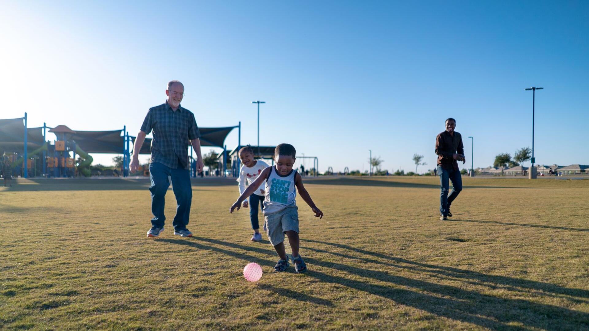 Dave, left, plays with Robert’s children, Sandra and Agape who call him Grandpa Dave. They are in a field and the children are kicking a ball. 