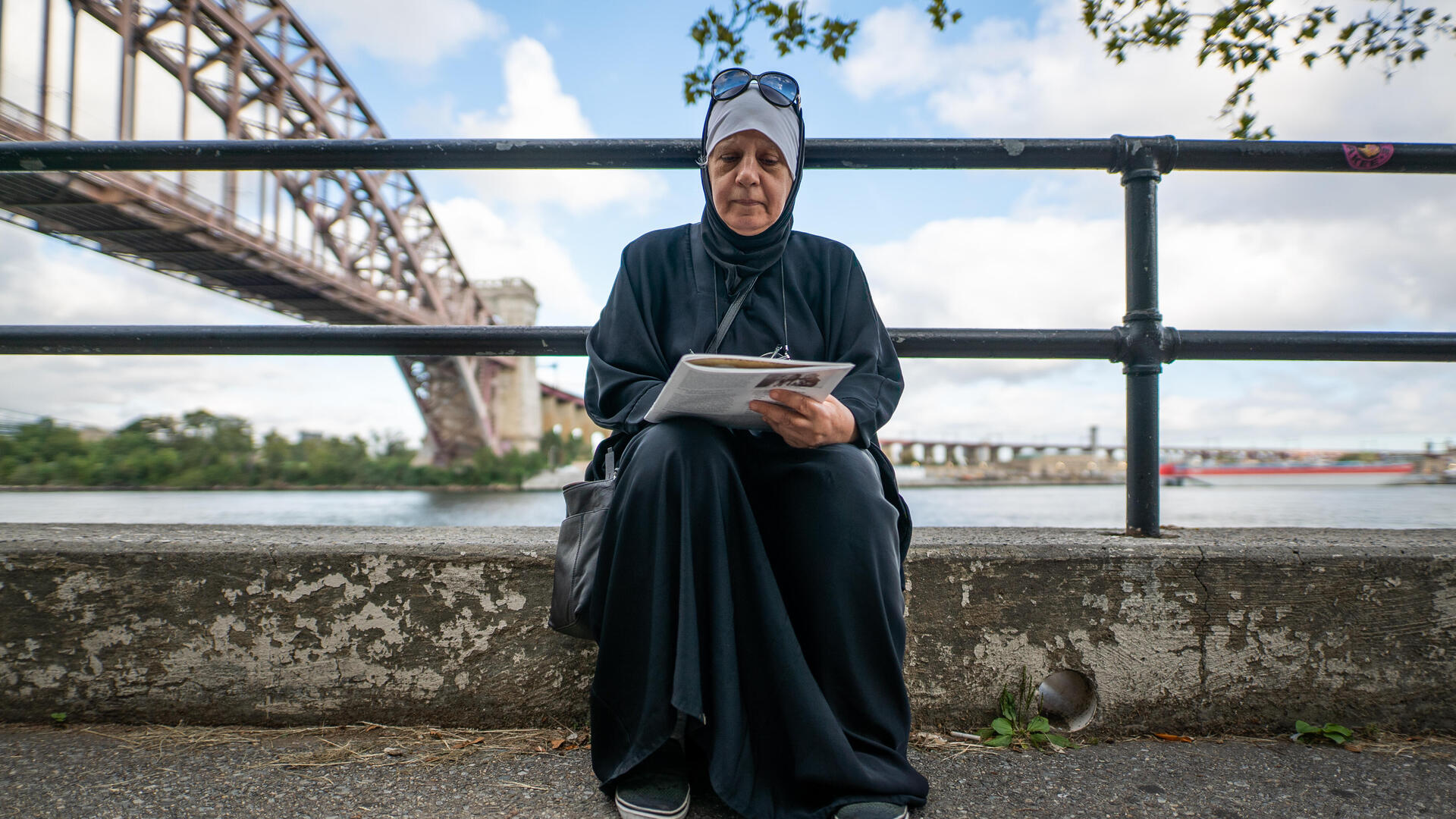 Maha sits on the curb next a river while looking at the book she uses to study for her citizenship. There is a bridge behind her in the distance. 