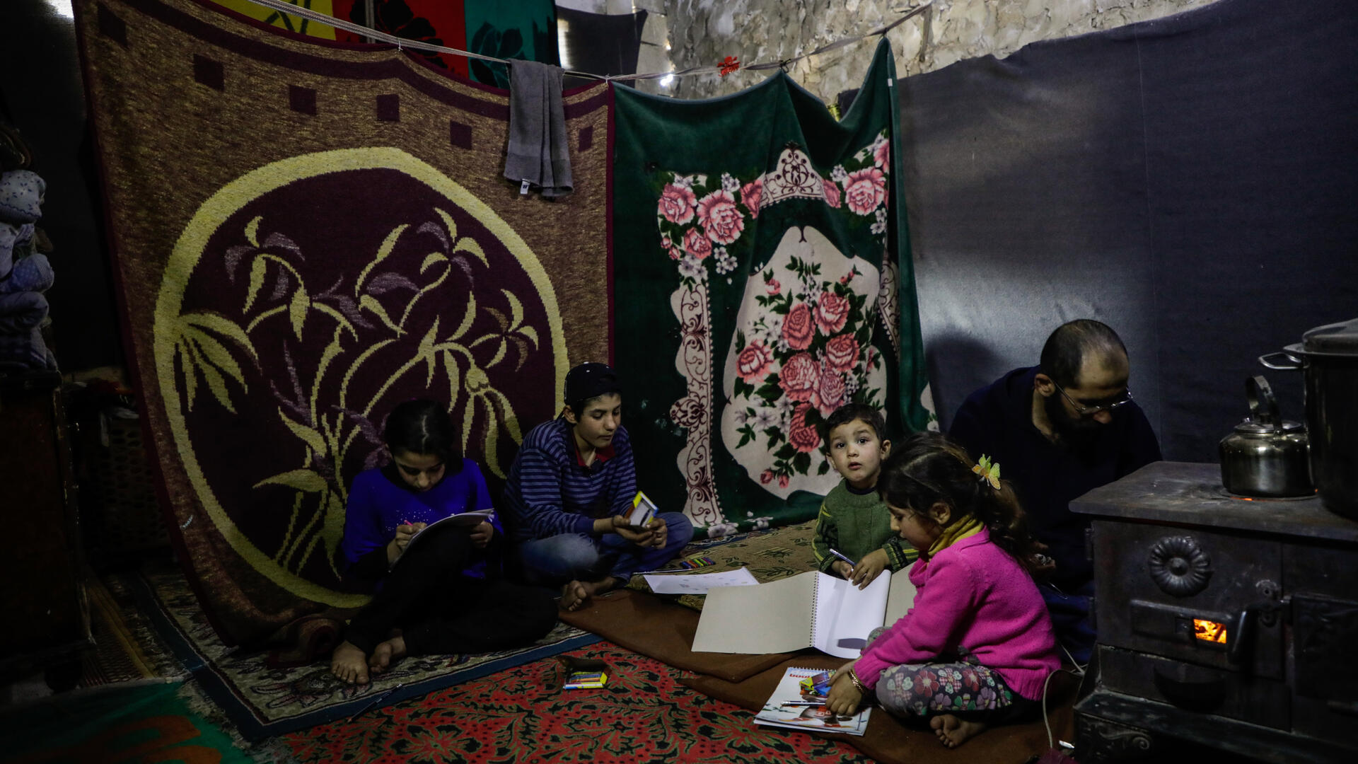 A displaced Syrian family living in an unfinished building with blankets for walls. The father tends to a small stove.