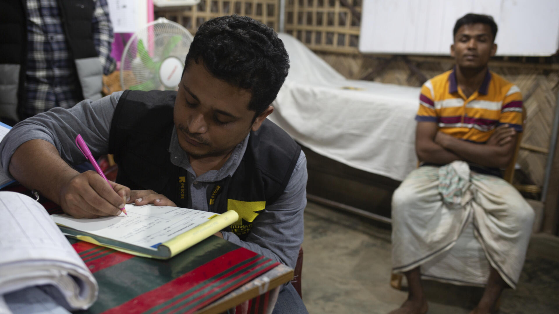 Dr. Mahmudul Hossain, wearing an IRC vest, sits at a desk writing notes. A patient sits behind him. 