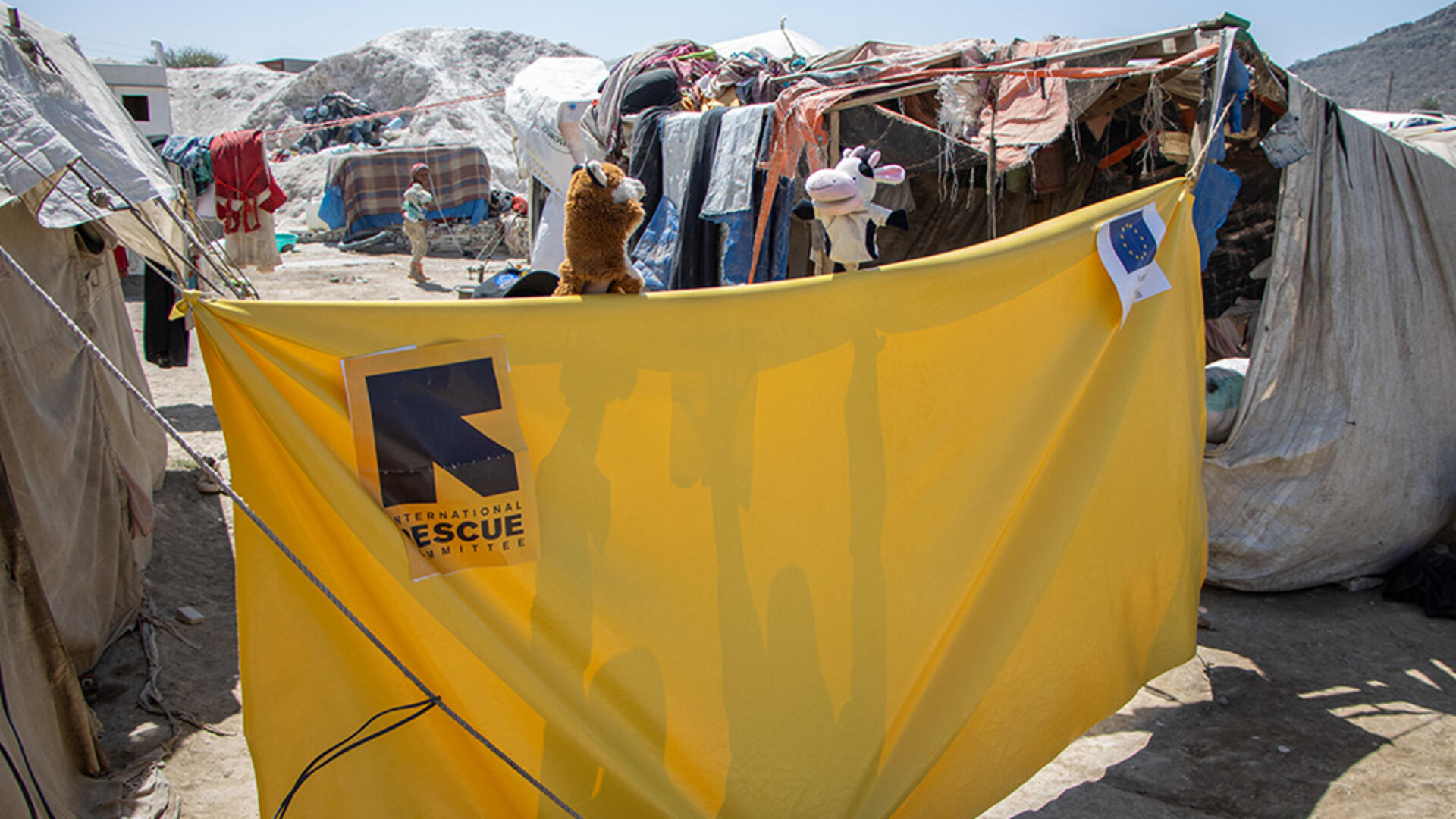 Outside in Al-Manshar camp in Yemen, a yellow tarp is set up between tents. Just above the tarp are a fox and cow puppet, part of the show to teach children about COVID-19. 