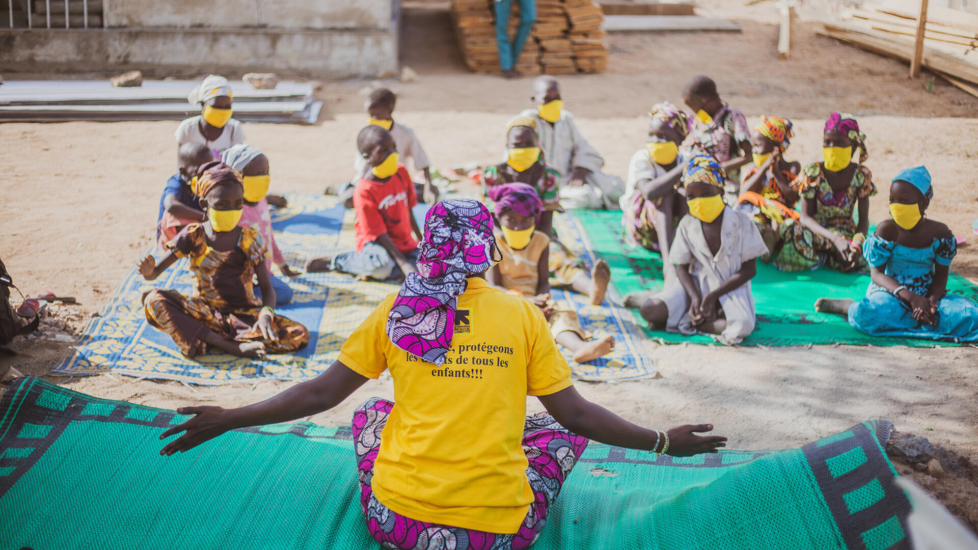 In addition to receiving academic support at this IRC Safe Healing and Learning Space in Cameroon, children are taught the importance of hygiene, as well as techniques to prevent the spread of COVID-19. A teacher with her arms spread out and seen from the back addresses a group of children seated socially distanced outdoors on a sheet, wearing masks.