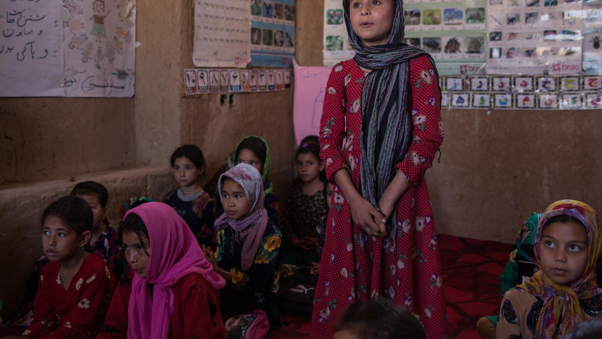 Children at an IRC Safe Healing and Learning Space in Afghanistan. A girl surrounded by her classmates stands up to speak while clasping her hands in an IRC safe healing and learning space in Afghanistan.