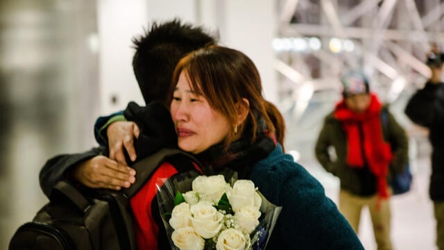 Crying mother hugs son while holding flowers at the airport.