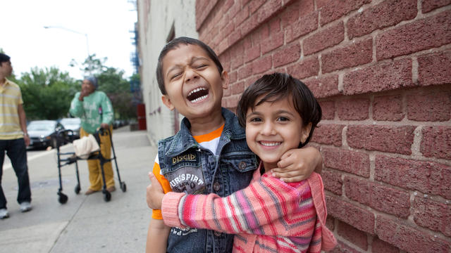 A young boy and girl stand outside in front of a brick wall, laughing and embracing.